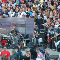 A punk rock band performs in front of a large, diverse crowd at an outdoor event reminiscent of Laulupidu. The band members play drums, guitar, and bass while people in the audience enthusiastically watch and sing along. The backdrop is filled with individuals holding song sheets, creating a lively, engaging atmosphere.