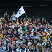 A large, diverse group of people sing in a choir, with some holding colorful folders and wearing vibrant costumes reminiscent of a Laulupidu festival. One person in the center holds a white flag with a black illustration. The dark background contrasts their enthusiasm and engagement, adding a touch of punk flair.