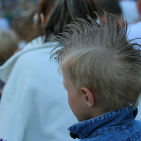 A child with spiky, blonde hair in a punk mohawk style is shown from behind, standing in a crowd at Laulupidu. The child is wearing a blue denim jacket. The background is blurred, featuring various people dressed in casual clothes, including someone in a white coat.