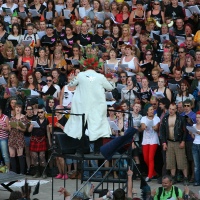 A conductor in a white coat and red spiked wig leads a large choir of diverse individuals at Laulupidu. The choir members are holding music sheets and appear engaged in singing. The scene is vibrant with a variety of punk outfits and hairstyles among the singers. The event takes place outdoors.