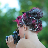 A person with dark pink and black hair styled in a punk updo decorated with chains and pins is holding a camera up to their face. They are wearing sunglasses, earrings, and a strapless top. The background is out of focus green foliage.