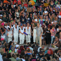 A large, colorful crowd is gathered outdoors, some dressed in bright costumes and wigs. The atmosphere echoes a spirited Laulupidu festival. At the center, a person in a gray shirt stands out against a group of people dressed in white. The lively scene has people holding papers, cheering, and some taking photos.