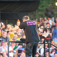 A man in a black t-shirt with pink text on the back stands at a podium, facing away from the camera. He appears to be conducting a lively outdoor crowd at Laulupidu, blending punk vibes with traditional charm. Spectators are under partly shaded areas, and greenery adorns the scene above.