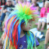 A child wearing a vibrant, punk-inspired multicolored wig with long spikes and dressed in a purple shirt stands among a seated crowd at Laulupidu outdoors. The wig features bright shades of green, yellow, orange, red, blue, and purple. The crowd in the background appears engaged and is slightly out of focus.
