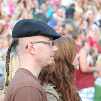 A man with a braided ponytail, glasses, and a black cap stands next to a woman with long brown hair in a crowd at an outdoor Laulupidu event. The background shows many people sitting on grass, creating a lively and social atmosphere with hints of punk fashion scattered throughout.