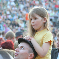A man wearing a hat carries a young girl on his shoulders at Laulupidu, a vibrant outdoor event. The girl, dressed in a yellow shirt, looks ahead with a thoughtful expression. In the background, the large crowd has a slightly punk vibe, enhancing the energy of the scene while remaining blurred to emphasize the foreground.