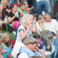 A young girl with blonde hair, wearing a striped shirt, sits on a man's shoulders in a crowded outdoor gathering reminiscent of Laulupidu. The man, donning a flat cap and white shirt with subtle punk flair, is surrounded by people sitting on grass, many engaged and holding snacks. The atmosphere is lively and social.