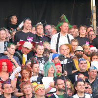 A large and diverse group of people are gathered closely together, many holding sheet music and singing. They are dressed in casual clothes, with some donning colorful wigs and punk accessories. Enthusiastic and focused, they stand against a dark backdrop, possibly indicating a stage setting reminiscent of Laulupidu.