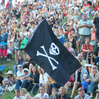 A large crowd sits on a grassy slope during Laulupidu, with people dressed in casual summer clothing. In the center, a black flag with a white skull and crossbones adds a punk flair. The audience appears engaged and diverse, featuring a mix of adults, children, and seniors.