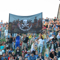 A diverse crowd is seated on a grassy slope at an outdoor gathering, reminiscent of Laulupidu. They are holding a large black flag with a white skull and crossbones design and the text "FOR ANARCHIST RESISTANCE." The punk-spirited crowd includes adults and children, all appearing engaged and attentive.