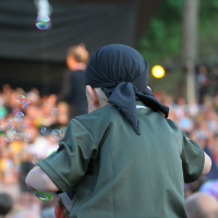 A person with a black headscarf and green shirt is seen from behind, sitting on someone's shoulders at an outdoor Laulupidu event. In the foreground, soap bubbles float around, and in the background, a large crowd of people can be observed amid a blurred punk performer on stage.