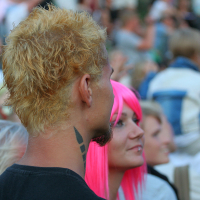 A man with bleach blonde hair and black geometric neck tattoos stands in a crowd at Punk Laulupidu, seen from behind. Beside him, a woman with a bright pink wig smiles while looking up. The background is blurred but filled with people wearing casual clothing.