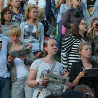 A diverse group of people are gathered outdoors, many holding newspapers. Some are standing, while others appear to be singing or reading, reminiscent of a Laulupidu celebration. The crowd includes men, women, and children in casual clothing. A few people in the background are taking photos and observing the scene.