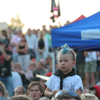 A young child with blue hair styled in a mohawk sits on someone's shoulders at an outdoor event. The child is wearing a white shirt and a black scarf. Surrounding the child is a large crowd of people under a blue canopy, suggesting a lively and energetic atmosphere reminiscent of Laulupidu.