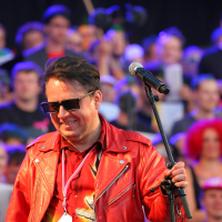 A man wearing a red leather jacket and sunglasses stands in front of a microphone on a stage. Smiling, he grips the microphone stand as if ready to deliver an electrifying punk performance. The crowd, slightly blurred, resembles a vibrant Laulupidu festival with colorful clothing, accessories, and signs.