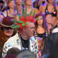 A person with spiked green and red hair, wearing a jacket with silver triangular patches and a large chain necklace, stands amidst the crowd at Punk Laulupidu. Another person with long dreadlocks and a maroon hat is near them. The background features a large group of people, some watching intently.