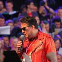 A man wearing a shiny red leather jacket and dark sunglasses holds a microphone, singing. He is surrounded by a large crowd of people, some holding papers. The background is colorful with blue, purple, and pink lighting. The lively and energetic mood evokes the spirit of Laulupidu mixed with punk vibes.