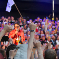 A performer in a red jacket sings on a stage, with a large crowd behind him. The audience in the foreground raises their hands, holding papers and wearing wristbands. The photo is vibrant with colorful lighting, and a flag is visible in the background, held by someone on stage at Laulupidu.
