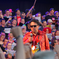 A man in a red leather jacket and sunglasses stands on stage with a microphone, channeling a punk vibe, surrounded by a large, diverse crowd holding papers and singing. Some audience members raise their fists and wear colorful accessories. The scene appears vibrant and lively, reminiscent of Laulupidu.