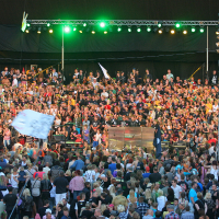 A large crowd at a lively outdoor concert at dusk. The stage, reminiscent of a punk rock scene, is lit with green and yellow lights, and musicians perform with a sea of people in colorful attire behind them. Scaffolding with speakers and lighting surrounds the area. Some attendees hold flags and banners, adding to the festive atmosphere.
