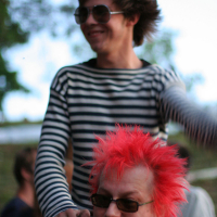 A young man in a striped shirt and sunglasses is sitting on the shoulders of an older man with vibrant spiked red hair and glasses, giving off a punk vibe. The older man smiles; both appear to be outdoors at Laulupidu with a blurred background of greenery and people.