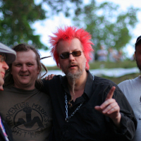 Four men are standing outdoors, three looking at the camera. The man in the center has bright pink, punk hair, sunglasses, and wears a black shirt with a chain necklace, pointing his finger. The man on his right wears a beige shirt with a face print. Trees and greenery are visible in the background.