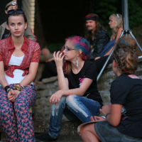 Three young people sit outdoors; one with a short red cardigan and printed pants, another with colorful pink and blue hair, wearing a black shirt and distressed jeans, giving off a punk vibe. A third faces away. In the background, a sitting man and woman are partially visible, with trees and a stone wall nearby at Laulupidu.