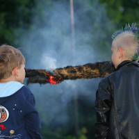 Two children are standing outside, facing away from the camera, watching a smoldering log suspended horizontally. One child is wearing a blue hoodie with a Snoopy graphic, and the other, sporting a Punk look with blonde hair styled in a blue and green mohawk, wears a black leather jacket. Smoke is visible around the log.
