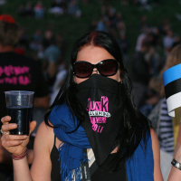 A person stands outdoors wearing sunglasses and a black face covering with pink text, exuding a punk vibe. They hold a drink in one hand and an Estonian flag in the other, likely at Laulupidu. The individual has long, dark hair and a blue scarf, with a crowd visible in the blurred background, suggesting a public event.