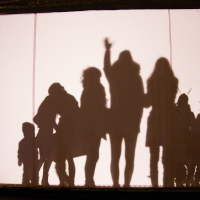 Silhouettes of several people on a backlit screen during the 2012 Rīga Staro festival, creating shadows of various poses, including one person waving and others standing together. The bright light behind the screen casts clear, dramatic outlines against the dark surroundings.