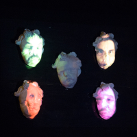 Five human faces sculpted in white material are arranged in a grid pattern. Each face is illuminated by colored lights, ranging from green, orange, purple, to pink, creating a dramatic and eerie effect against a dark background. This stunning display was featured at the 2012 Staro Rīga festival.