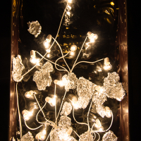 A display features lit light bulbs connected by white wires, surrounded by crumpled sheets of aluminum foil. The bulbs and foil are arranged artistically inside a dark rectangular frame, giving the effect of glowing elements amidst a metallic, reflective backdrop—reminiscent of Rīga's Staro 2012 festival.