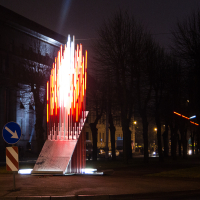 A night scene depicts a Staro Rīga 2012 modern art installation with numerous vertical light rods, most of which are illuminated in red, forming a flame-like shape. Nearby, leafless trees and street lamps light up the background. Directional road signs are present on the left, guiding traffic.