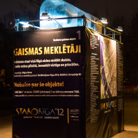 A rectangular exhibition booth is seen at night, lit by overhead spotlights. The booth features posters with detailed text in Latvian, including large titles, event details, and a map. The text "GAISMAS MEKLĒTĀJI" is prominently displayed as part of the 2012 "Staro Rīga" festival.
