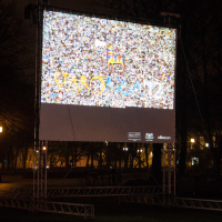 In a dimly lit park with trees in the background, an outdoor movie screen is set up. The screen displays a colorful and pixelated image with "RANDOM" visible in the center, reminiscent of Staro Rīga 2012. Several logos adorn the screen's frame. The surrounding area is dark, save for a few glowing park lights.
