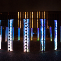 A nighttime photo showcases a circular art installation composed of illuminated fabric panels, each displaying vertical blue and white light patterns. In the background, a building adorned with vertical yellow lights enhances the vibrant ambiance of the scene, reminiscent of Rīga's Staro festival in 2012.
