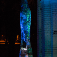 A tall, abstract ice sculpture illuminated by blue and green lights stands between two white brick archways in Rīga during the 2012 Staro festival. The sculpture has a smooth, elongated form with a texture that glitters under the light. The background is dark, enhancing the vibrant illumination of the ice.