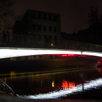 A bridge with cylindrical railings arches over a calm body of water at night. Illuminated by a bright white light along its length, the reflection shines in the water below. A dark building stands silhouetted in the background, with red lights reflecting in the water as well—capturing the essence of Staro Rīga 2012.