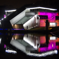 Modern building with tilted roof and large glass sections stands illuminated at night by white, purple, and blue lights. Reflecting clearly in the calm water of a nearby pond, this architectural gem was part of the 2012 Staro Rīga festival. The surrounding area is dimly lit with trees and grass barely visible in the background.