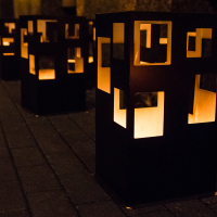 A row of black metal lanterns with cut-out square patterns, each containing a lit candle, placed on a cobblestone walkway in Rīga's Staro 2012 festival. The warm glow from the lanterns creates a cozy and inviting atmosphere, illuminating the path.