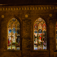A brick wall of a church in Rīga is adorned with four large, illuminated stained glass windows at night. Each window contains detailed, colorful religious scenes with figures, intricate designs, and ornate patterns highlighting significant biblical narratives. The display first captivated the public during Staro 2012.
