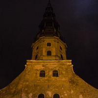 A tall, gothic-style brick tower at night, illuminated by yellow lights. The tower narrows as it rises, with multiple tiers, each becoming progressively narrower. The 2012 Rīga Staro installation beautifully contrasts against the night sky in the background.