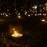 The image shows numerous small tea light candles arranged on grassy ground at night, creating a serene and somber atmosphere. The candles emit a warm glow, illuminating the immediate area, while the background is dark with blurred lights from distant buildings. This scene is reminiscent of Rīga's Staro festival in 2012.