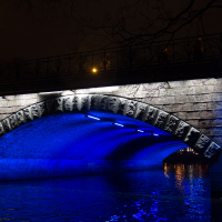 A nighttime view of a stone bridge in Rīga illuminated with blue lights from the 2012 Staro festival, reflecting off the calm water below. The bridge has ornate railings and is adorned with decorative stonework. Streetlights above cast a warm, soft glow, contrasting with the cool blue lights underneath. Skeletal tree branches are visible along the top edge.