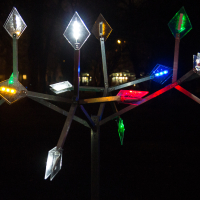 A nighttime photo of Staro Rīga's 2012 lighted art installation featuring a tree-like structure with multiple diamond-shaped lights in various colors, including white, red, green, and blue. The dark background highlights the illuminated elements, creating a striking visual contrast.