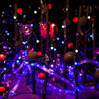 A vibrant and intricate display of multicolored lights, featuring a mix of red, blue, and white bulbs reminiscent of 2012's Staro Rīga festival. The setup includes black poles adorned with spherical decorations and illuminated tubes, creating a visually dynamic, almost whimsical scene with a warm, glowing ambiance.