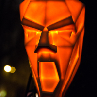 A glowing, orange, geometric mask with exaggerated facial features, including a wide open mouth and angular eyes, set against a dark, blurred background at the 2012 Staro Rīga festival. The mask emits a sharp, illuminating light, highlighting its intricate, polygonal design.