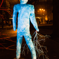 A blue, human-shaped sculpture stands outdoors at night during Staro Rīga 2012, holding a bundle of branches. The figure is illuminated by several lights and appears wrapped in blue material. A red light illuminates the nearby branches, creating a stark contrast with the surrounding dark trees and distant streetlights.