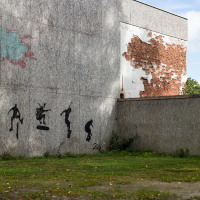 Silhouetted street art of figures in motion adorns a weathered wall in Saaremaa, contrasting with a patch of exposed brick under fading graffiti, while nature quietly reclaims the surrounding