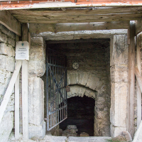An old stone entrance in Saaremaa, secured with a metal gate, leading into a dark, mysterious underground space, complemented by a warning sign and framed by wooden beams, evoking a